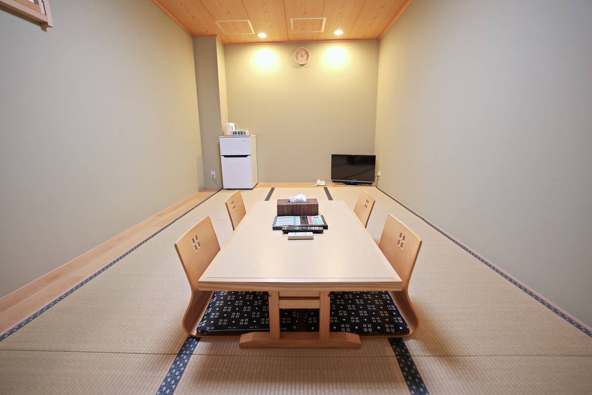 A tatami mat room with a wooden table, chairs, a television, and a refrigerator.