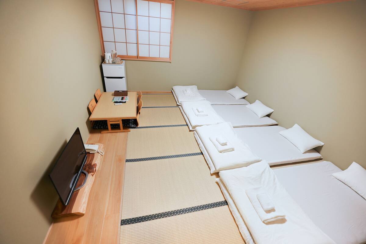 A room with a wooden desk, a laptop, a small table with a cup and a book, and a bed with white bedding.