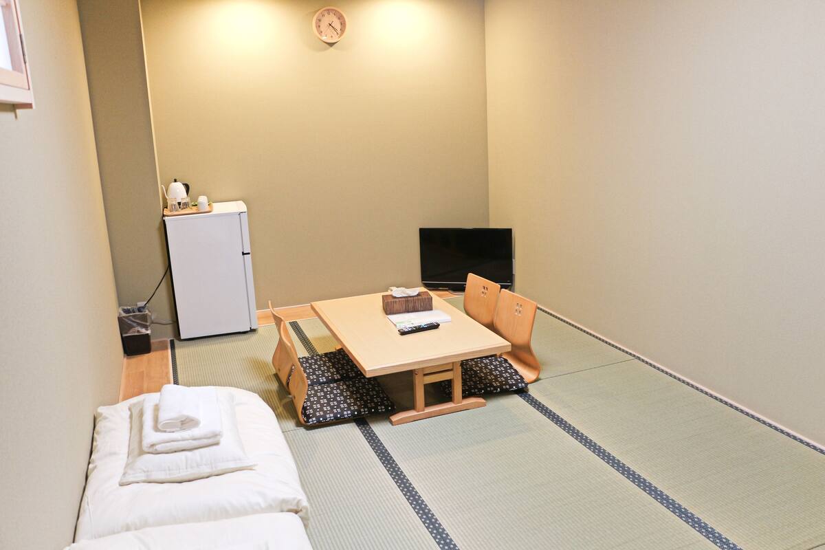 A room with a bed, a small table with a chair, a television, and a refrigerator.