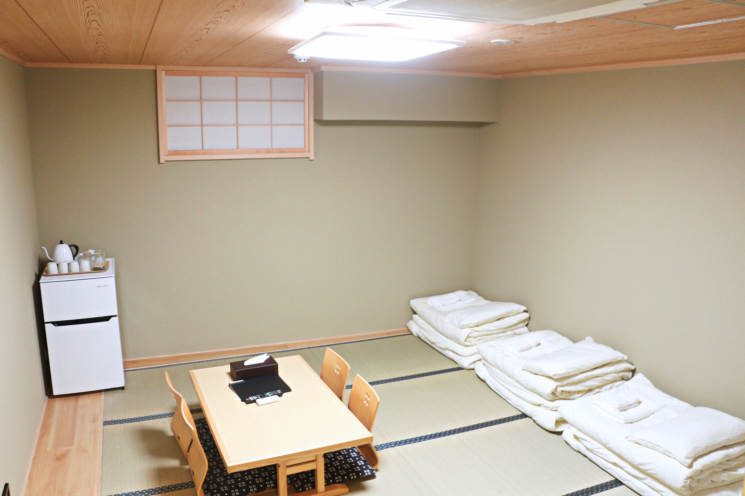 Een kamer met tatami vloerbedekking, een lage tafel met stoelen, een kleine koelkast en netjes opgestapelde dekens.