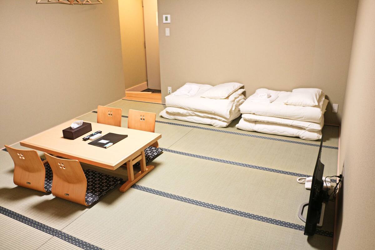 A hotel room with tatami flooring, a wooden table with chairs, and neatly folded towels on a shelf.
