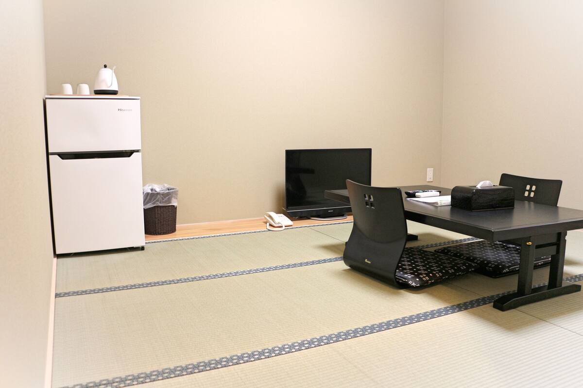 A room with a dining table, chairs, a black TV, a white refrigerator, and a trash bin.
