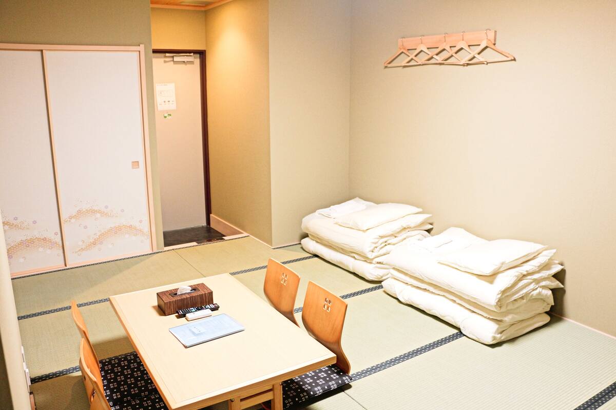 A traditional Japanese room with tatami flooring, a low table with a cushion, a bed with neatly folded bedding, and a wooden shelf with hangers.