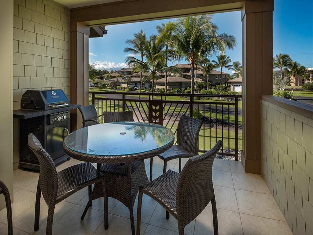 Een overdekt terras met een ronde glazen tafel, rieten stoelen, een barbecue en uitzicht op palmbomen en huizen.