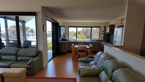 A modern living room with a sofa, a dining table, and a kitchen area with cabinets and a refrigerator. - "LAZY DAYZ" Currarong beach home.   50m from unspoilt beaches and walking tracks (Currarong)