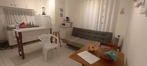 A compact kitchen with a refrigerator, microwave, and stove. There is a dining table with a chair and a small stool. A sofa is placed against the wall, and a window with curtains is visible.