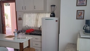 A compact kitchen with white cabinets, a refrigerator, a blender, a gas stove, and a dining area with a table and chairs. - Studio Morro das Pedras (Florianópolis)