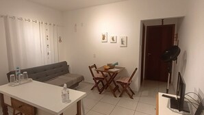 A small, well-lit room with a white table, a grey sofa, and a dining table with chairs. There is a microwave on the left, a door leading to another room, and a framed picture on the wall. - Studio Morro das Pedras (Florianópolis)