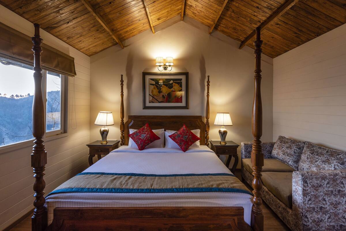 A four-poster bed with red and white bedding, two bedside lamps, a framed picture above the bed, and a patterned sofa to the right.