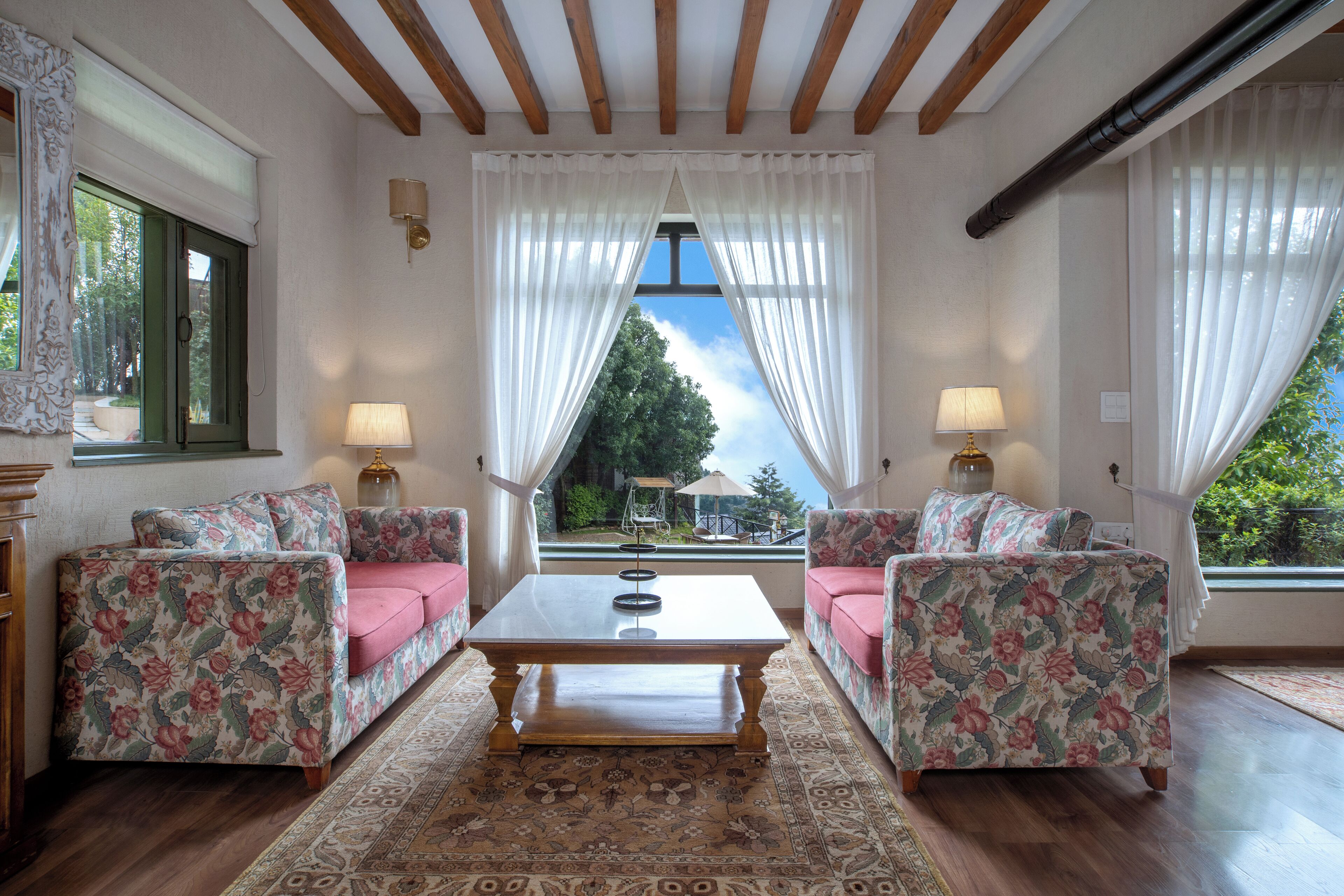 Una habitación con dos sofás de estampado floral, una mesa de centro y vistas a través de una ventana con cortinas blancas.
