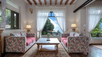 Una habitación con dos sofás de estampado floral, una mesa de centro y vistas a través de una ventana con cortinas blancas.