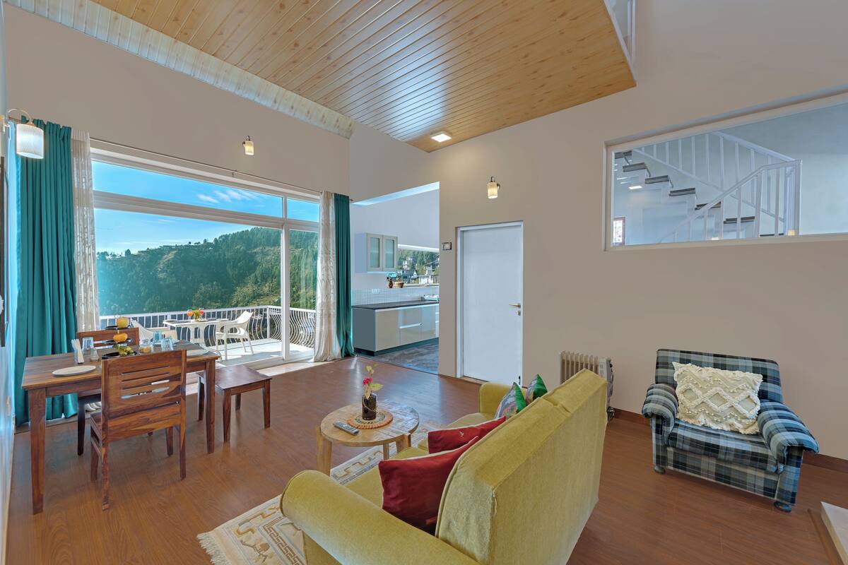 A spacious living area with a wooden ceiling, a dining table with chairs, a sofa, a chair with a cushion, and a view of a kitchen and balcony.