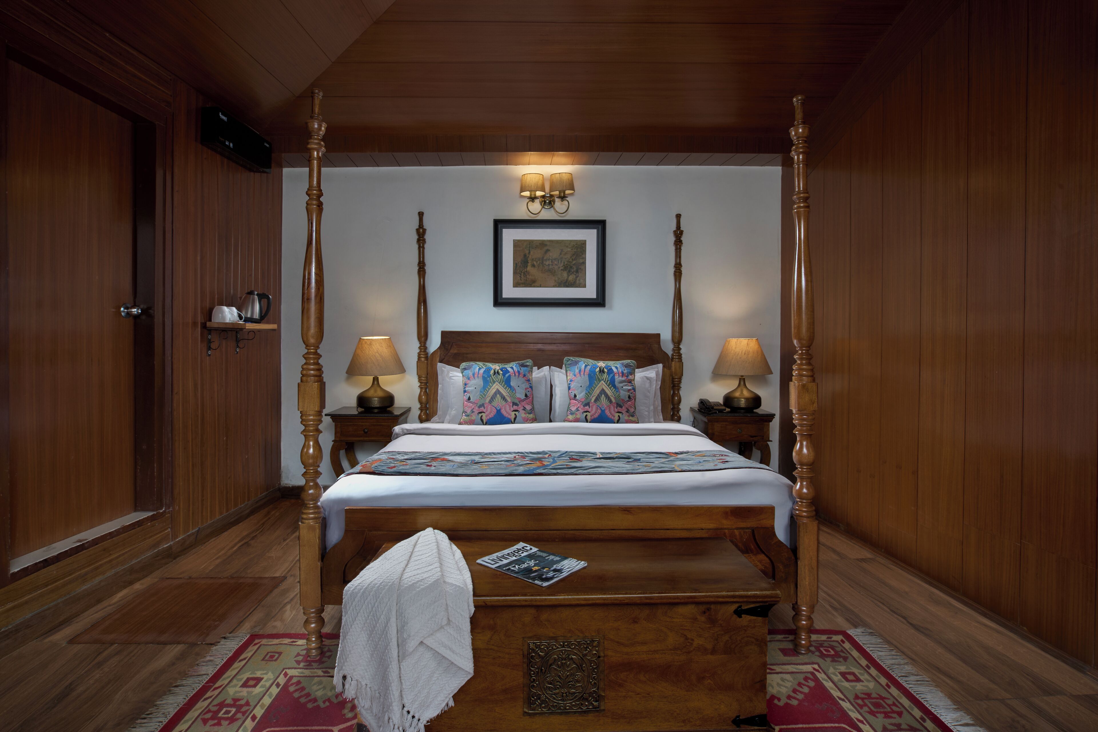Un lit à baldaquin avec une structure en bois et un couvre-lit blanc, flanqué de deux tables de chevet avec des lampes, une image encadrée au-dessus du lit et une armoire en bois sur le côté gauche.