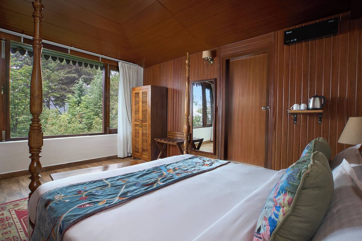 A bedroom with a large bed, a wooden wardrobe, a small table, and a window with a view of greenery.