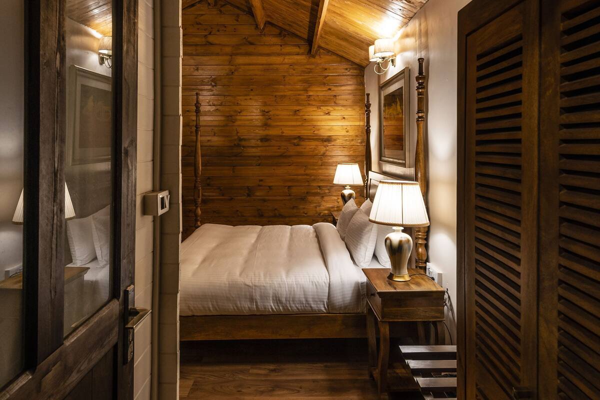 A narrow hallway leading to a wooden-paneled bedroom with a bed, bedside lamps, and a wooden door.