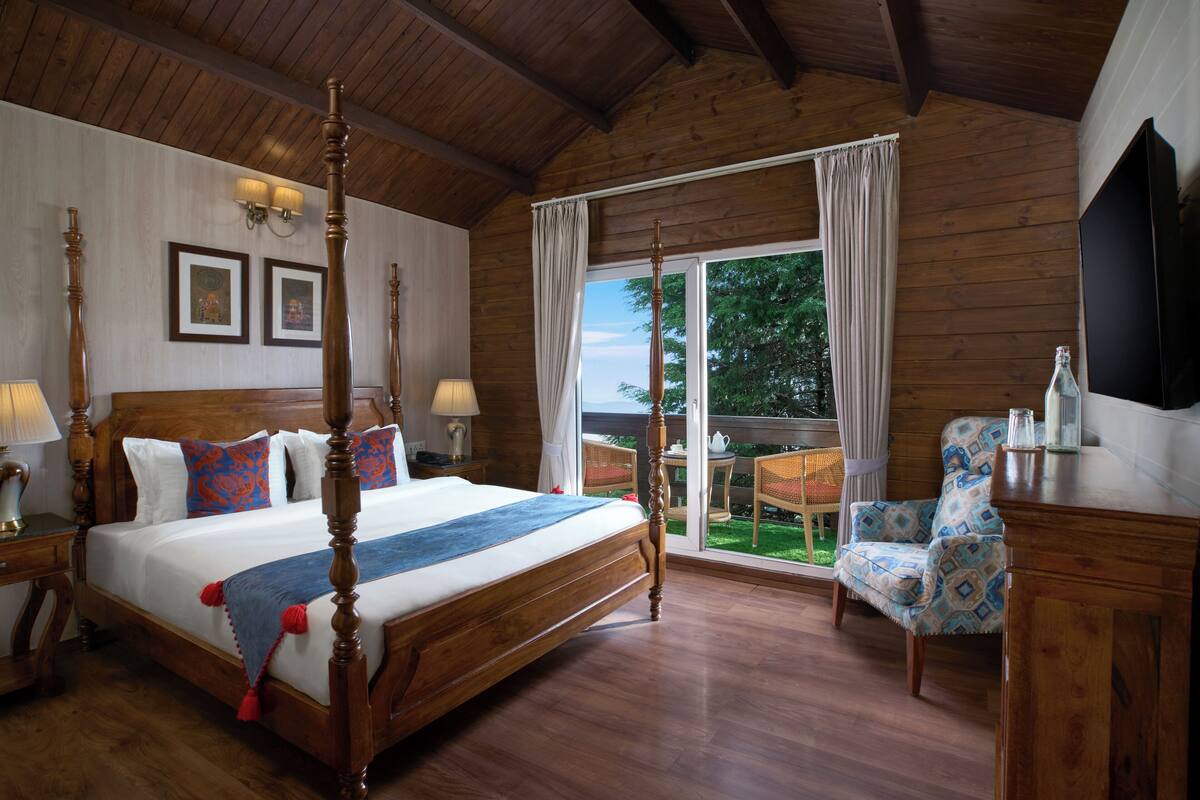 A four-poster bed with a wooden headboard, two bedside tables with lamps, a chair, and a television on a wooden cabinet.