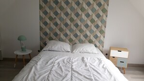 Una cama bien hecha con ropa blanca, un papel tapiz estampado en la pared de fondo, una mesita de noche con una lámpara y un atril con un cajón.