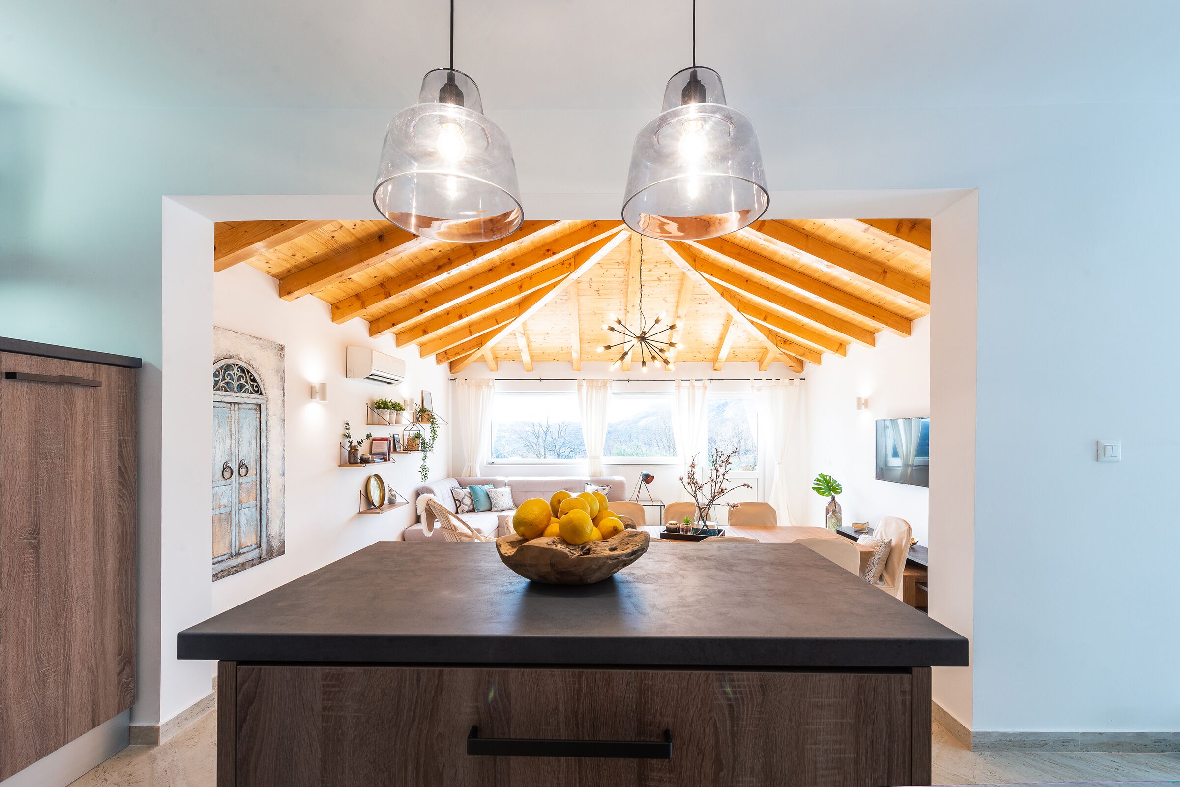 A spacious kitchen with a large island, wooden ceiling beams, pendant lights, and a bowl of lemons on the island.