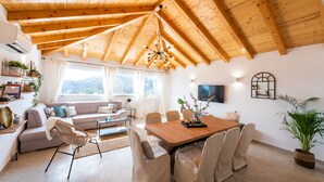 Een ruime woonkamer met houten plafond, een eettafel met stoelen, een bank en een televisie aan de muur.