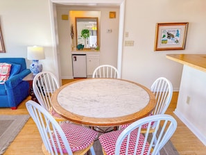 A round dining table with white chairs, a blue sofa, and a small kitchen area in the background.