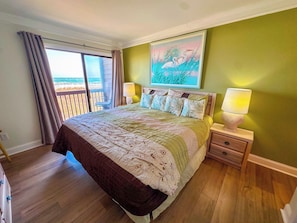 A bedroom with a bed, a nightstand, a lamp, a painting, and a view of the beach.