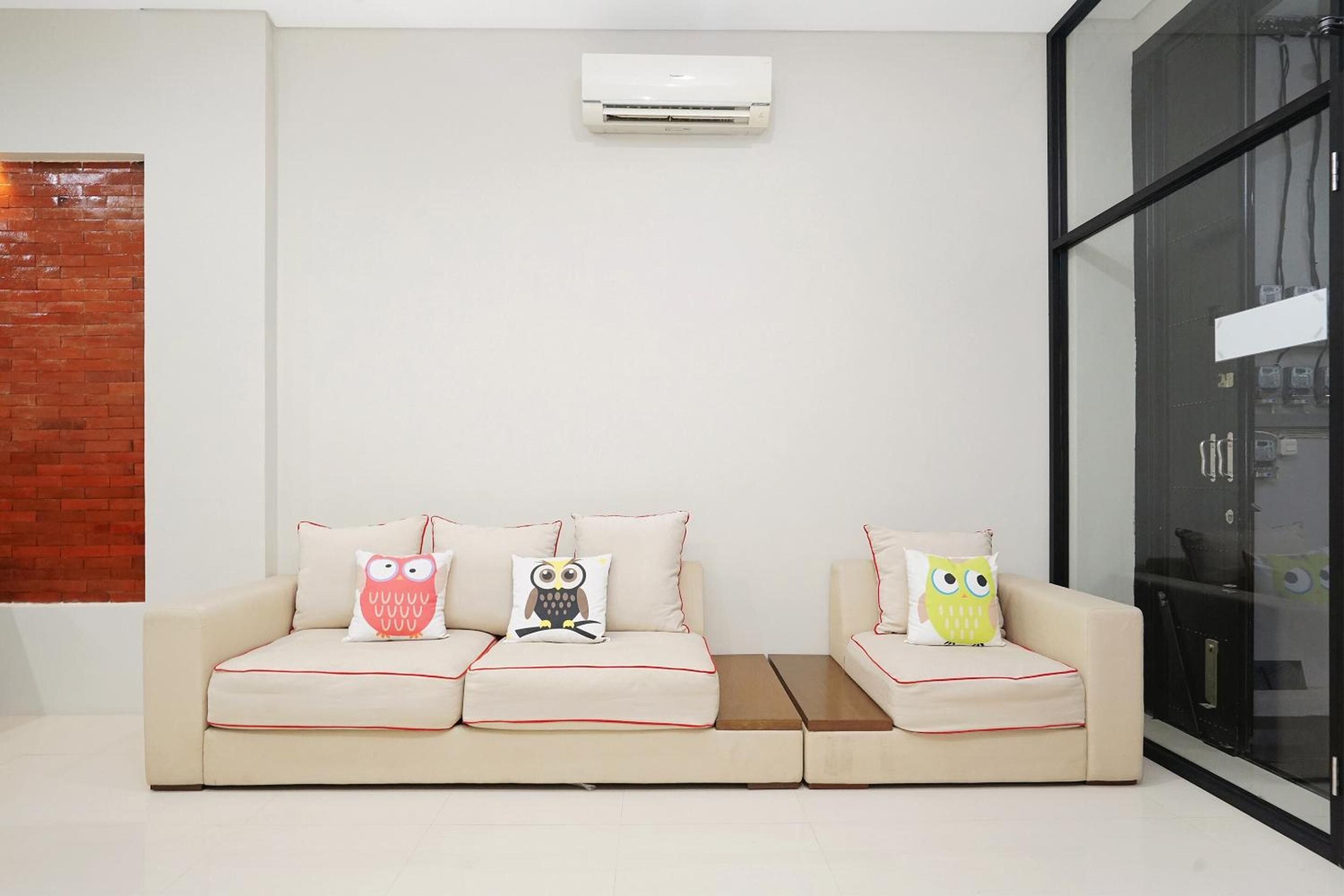 A modern living room with a beige sofa, three owl-themed cushions, and a wall-mounted air conditioner.