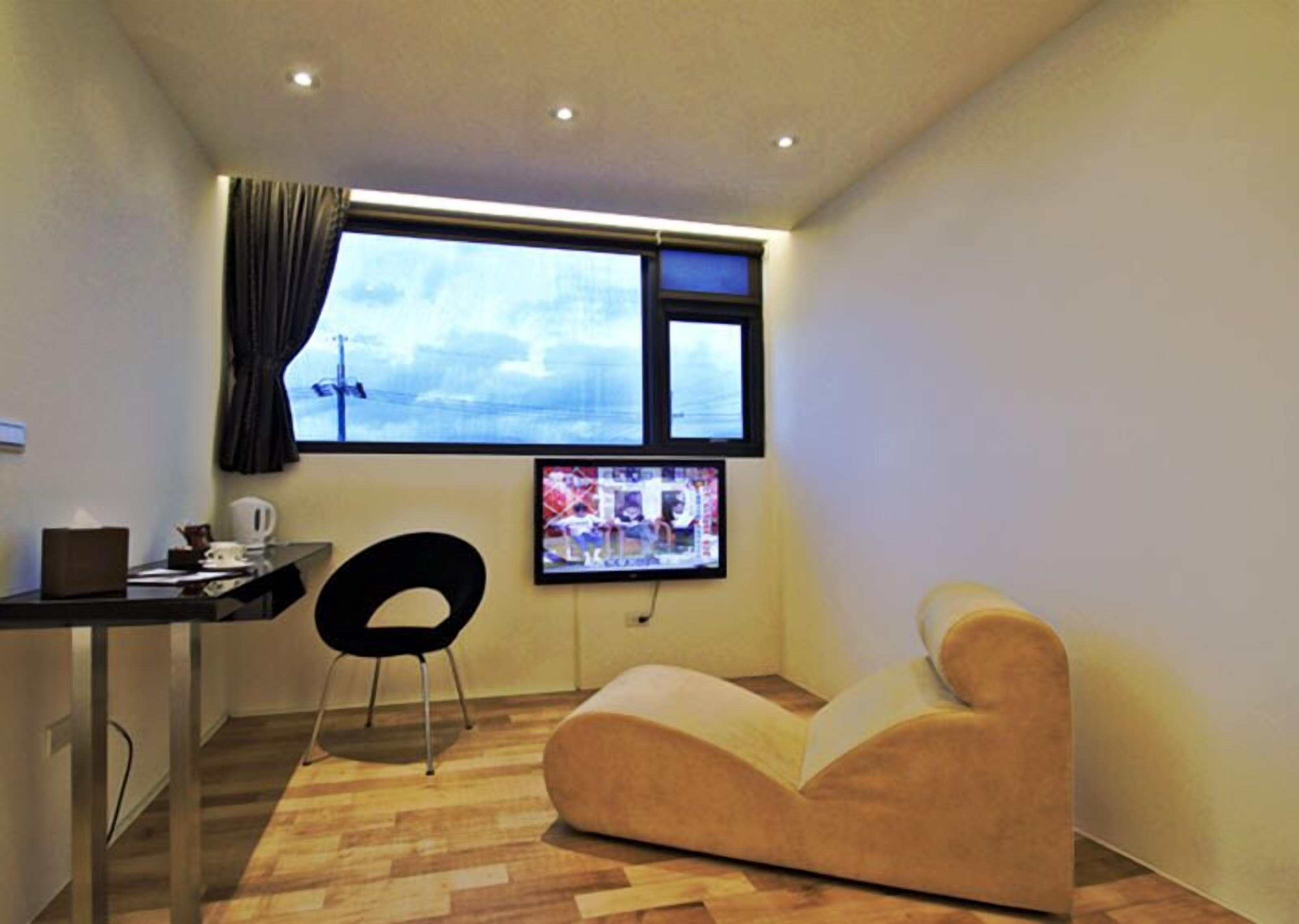 Ein kleiner, moderner Raum mit Schreibtisch, schwarzem Sessel, Sitzsack, Fernseher und einem Fenster mit Vorhängen.