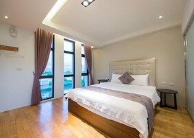 A modern hotel room with a large bed, a nightstand, and a view of the outdoors through large windows.