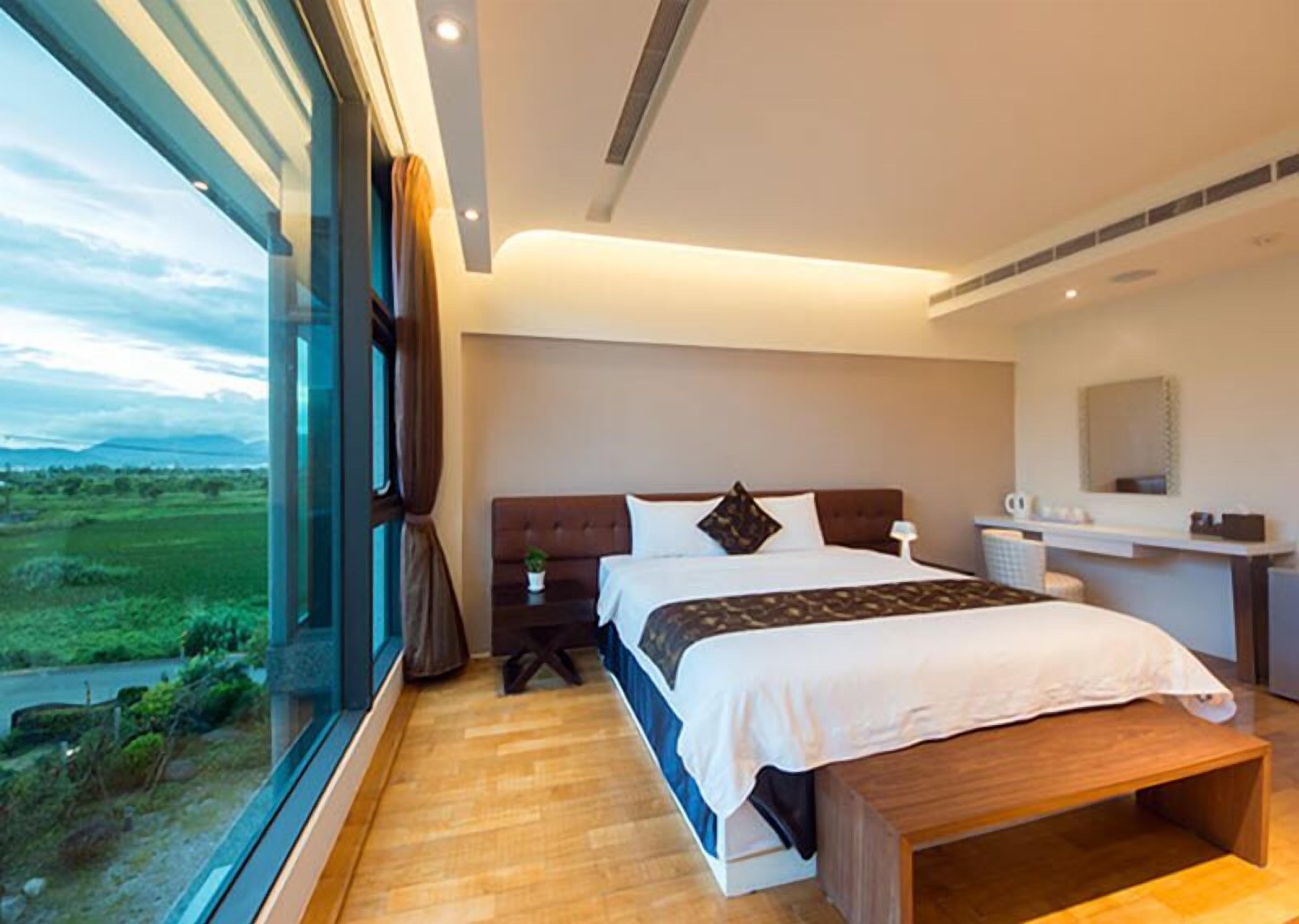 A modern bedroom with a large bed, a bedside table, a nightstand, a window with curtains, and a view of a green landscape.