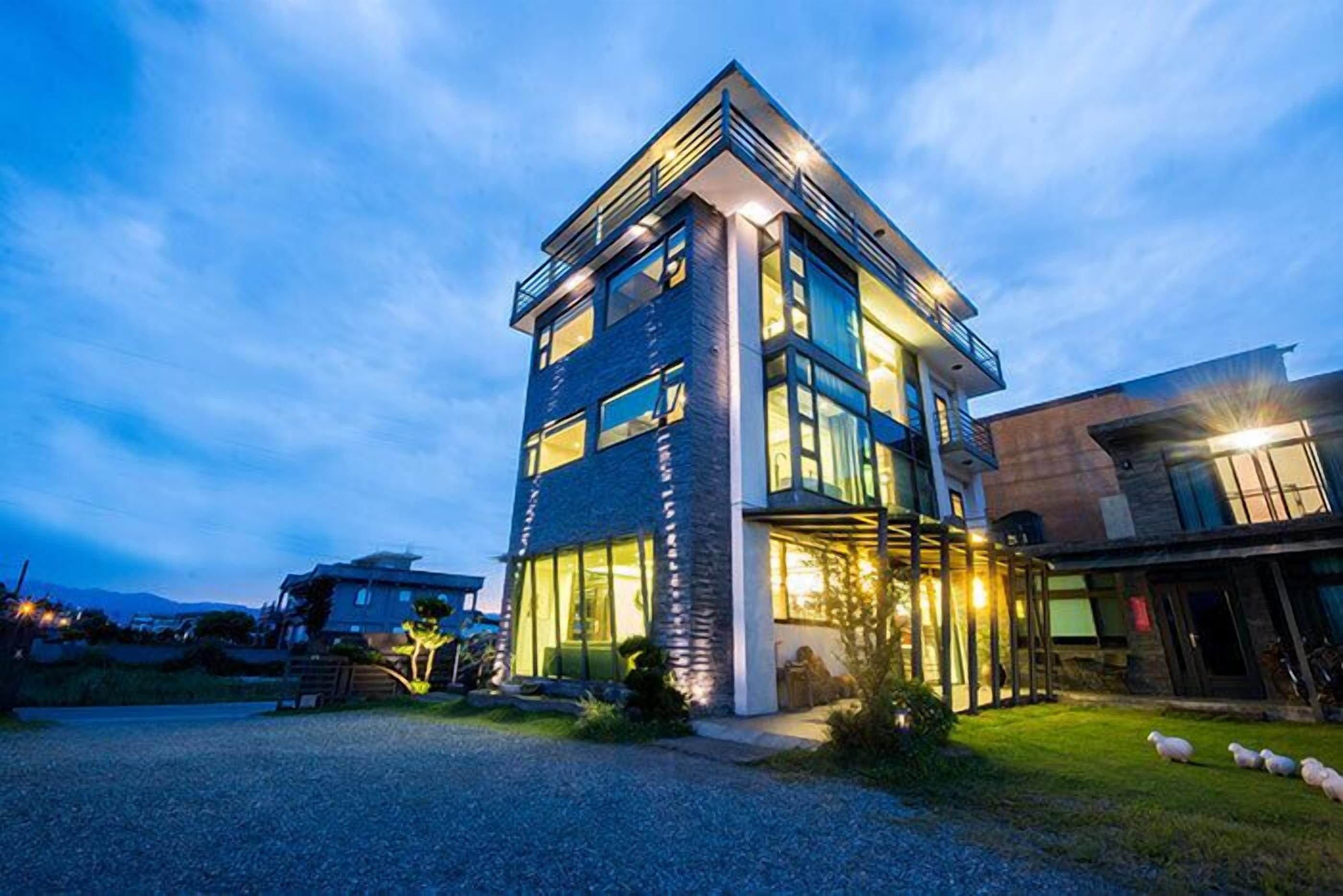 A modern two-story house with large windows and a balcony, surrounded by a well-maintained lawn and a gravel driveway.