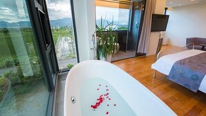 Een moderne hotelkamer met een groot bad vol rozenblaadjes, een bed met een blauw beddegoed met patroon, een flatscreen televisie en uitzicht op een groen landschap door grote ramen.
