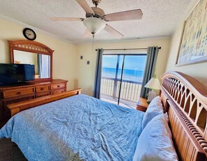 A bedroom with a bed, a dresser, a ceiling fan, a window with a view of the beach, and a clock on the wall.