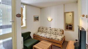 Et lille, hyggeligt værelse med sofa, stol, lille bord, fjernsyn og spejl.
