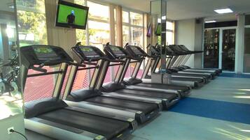Varias cintas de correr en un gimnasio con un televisor en la pared, ventanas amplias y piso cubierto de moqueta azul.