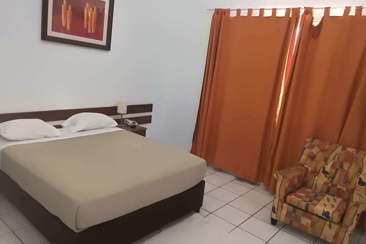 Una habitación de hotel con cama, mesita de noche, teléfono, silla y cortinas naranjas.
