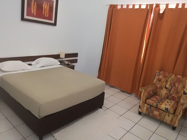 Una habitación de hotel con cama, mesita de noche, teléfono, silla y cortinas naranjas.