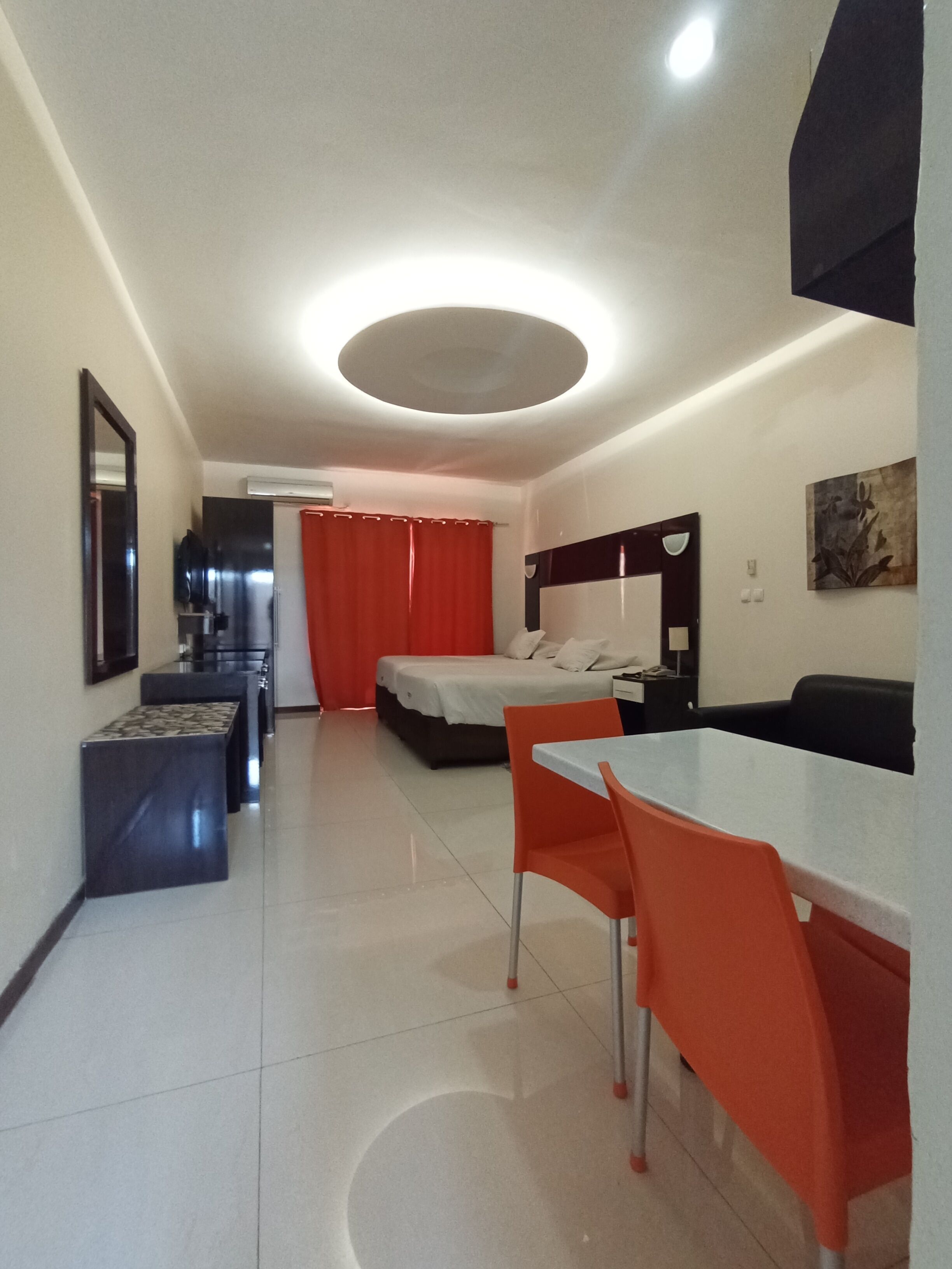 A modern hotel room with a bed, a dining table with orange chairs, a small kitchenette, and a bathroom with a shower curtain.
