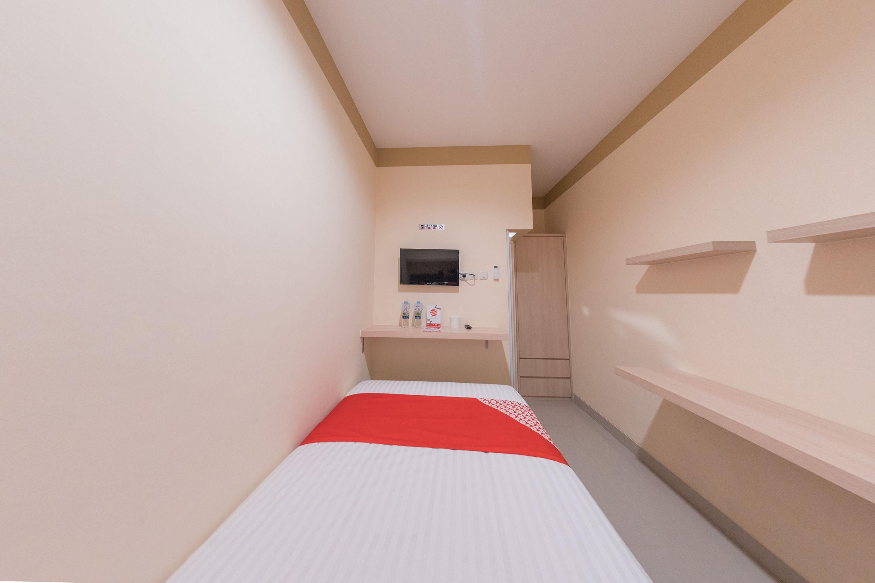 A narrow hotel corridor with a single bed, a television, and a small shelf with toiletries.