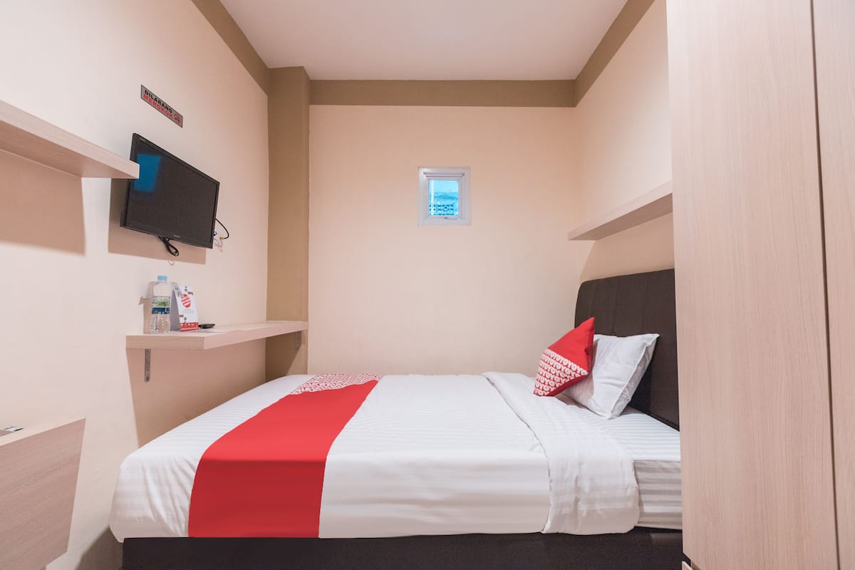 A hotel room with a bed, a television mounted on the wall, a small window, and a shelf with a bottle and a box.