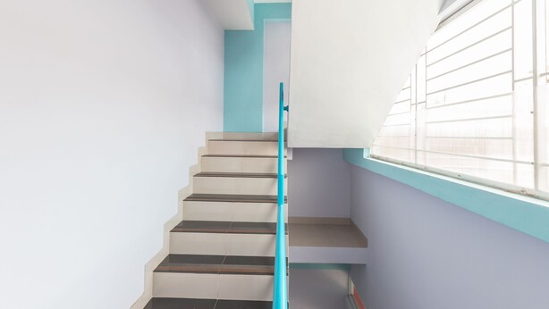 A staircase with a blue handrail, white walls, and a window with blinds. - Hotel O Balcon Biru (Jakarta)