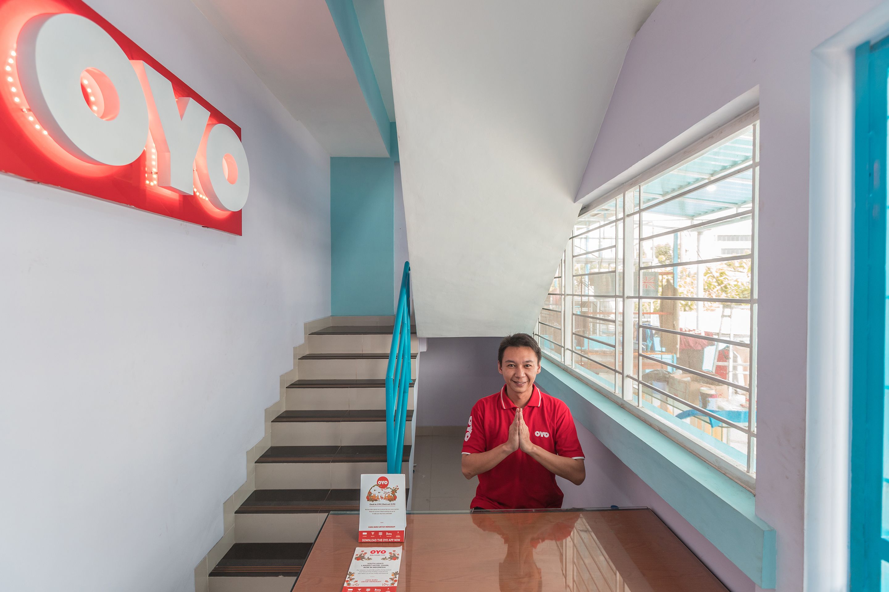 Une personne portant un t-shirt rouge se tient dans une pièce qui contient un escalier, une fenêtre donnant sur un paysage et un panneau avec les lettres « OYO ».