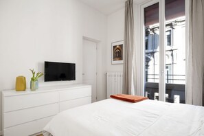 Un dormitorio con cama, televisor sobre un tocador blanco, ventana con vista a edificios y una puerta.