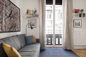 A modern living room with a grey sofa, a large cityscape painting, and a view of a building through a glass door. - The apartment is located in the historic center just three minutes from the fountain of Neptune and a short walk from all the most important artistic and culinary destinations of the city.In this neighborhood you can breathe a ville lumière atmosphere (Bologna)