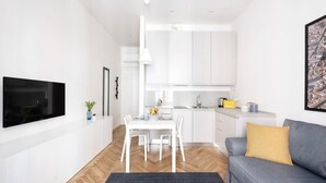 A modern living room with a flat-screen TV, a gray sofa with a mustard pillow, a dining table with chairs, and a kitchen area with white cabinets and a hanging light. - The apartment is located in the historic center just three minutes from the fountain of Neptune and a short walk from all the most important artistic and culinary destinations of the city.In this neighborhood you can breathe a ville lumière atmosphere (Bologna)