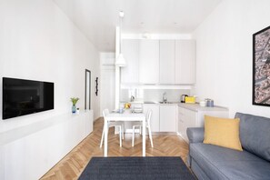 A modern living room with a flat-screen TV, a gray sofa with a mustard pillow, a dining table with chairs, and a kitchen area with white cabinets and a hanging light.