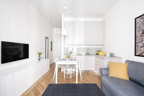 Ein modernes Wohnzimmer mit Flachbildfernseher, einem grauen Sofa mit Senfgelb-Kissen, einem Esstisch mit Stühlen und einer Küchenzeile mit weißen Schränken und einer Pendelleuchte.