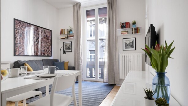 A modern living room with a dining table, chairs, a sofa, a large painting, a bookshelf, a radiator, and a window with curtains. - The apartment is located in the historic center just three minutes from the fountain of Neptune and a short walk from all the most important artistic and culinary destinations of the city.In this neighborhood you can breathe a ville lumière atmosphere (Bologna)