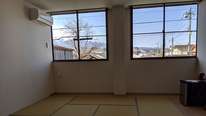 A room with tatami mat flooring, two large windows with a view of buildings and power lines, and a small heater on a table. - Machi no Hotel (Fujiyoshida)