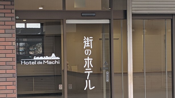 The entrance to Hotel de MACHI with a glass door and a sign in Japanese. - Machi no Hotel (Fujiyoshida)