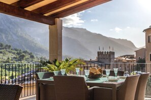 Un tavolo apparecchiato all'aperto con sedie in vimini, un tavolo in vetro e vista sulle montagne e un castello.
